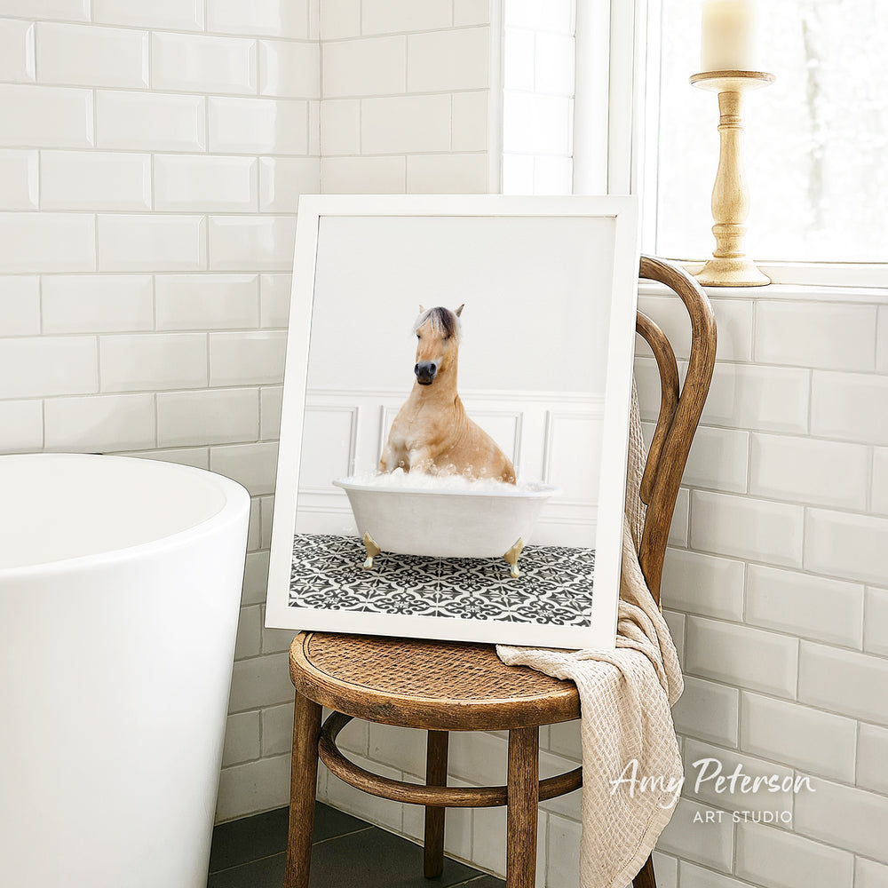 A framed image of a horse sitting in a bathtub, surrounded by a patterned tile design, is displayed on a wooden chair in a bathroom setting.