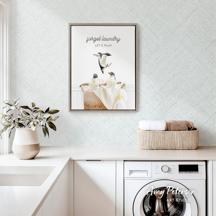 a framed poster of penguins sitting in a basket, hanging on a wall above a white countertop with a plant and a basket of towels.
