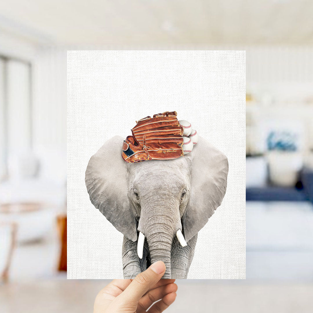 A hand holds up a framed image of an elephant with baseball gloves on its head, creating a playful and whimsical scene.