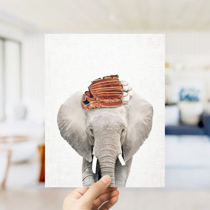 A hand holds up a framed image of an elephant with baseball gloves on its head, creating a playful and whimsical scene.