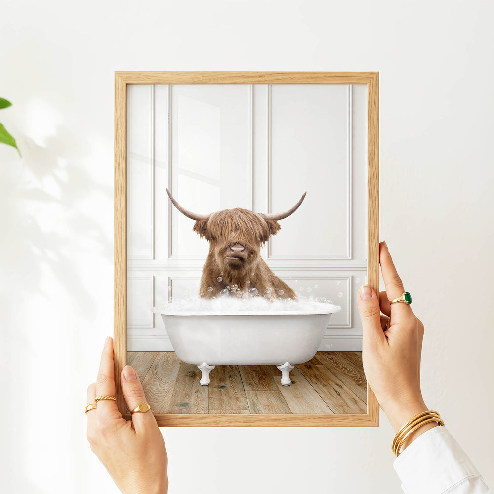 A person's hands are holding a picture frame containing an image of a Highland cow sitting in a bathtub filled with bubbles.