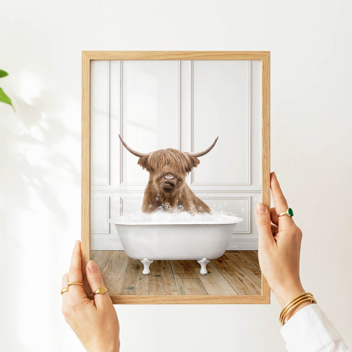 A person's hands are holding a picture frame containing an image of a Highland cow sitting in a bathtub filled with bubbles.