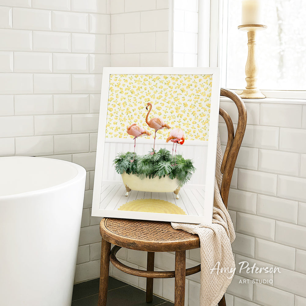 A framed print of a vibrant, colorful painting depicting a group of flamingos standing in a bathtub filled with green plants, placed on a wooden chair in a bathroom setting.