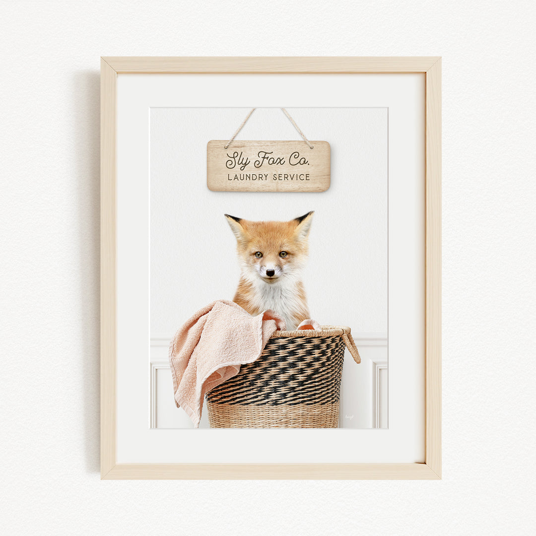A framed photograph of a young fox sitting in a woven laundry basket, with a sign above it that reads "Laundry Service".