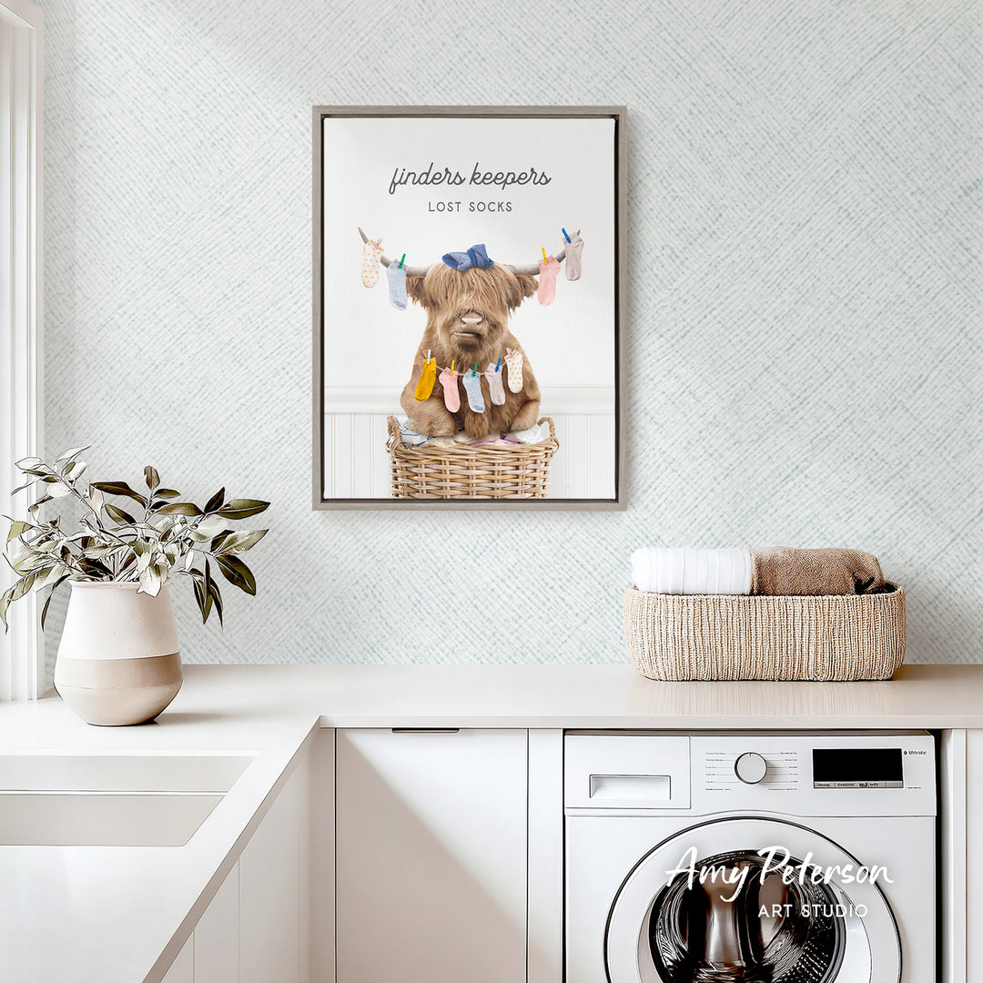 a framed poster of a Highland cow hanging socks on a clothesline in a basket, placed above a white kitchen counter with a potted plant and a basket of towels.