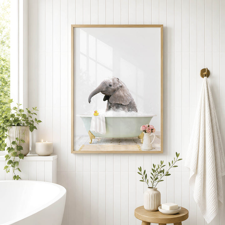 A framed photograph of an elephant sitting in a bathtub, surrounded by flowers and plants, is displayed on a white wall in a bathroom.