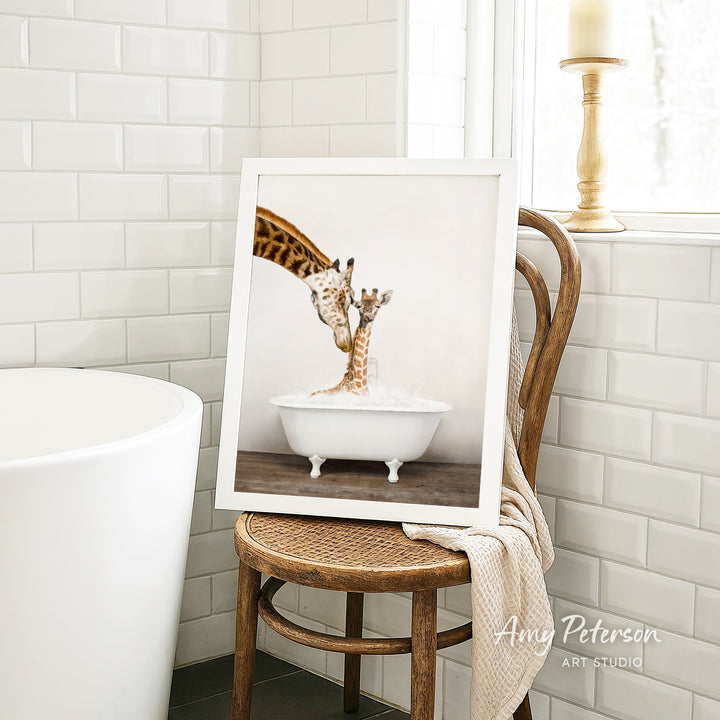 A framed image of a giraffe standing in a bathtub, with another giraffe inside the tub, is displayed on a wooden chair in a bathroom setting.