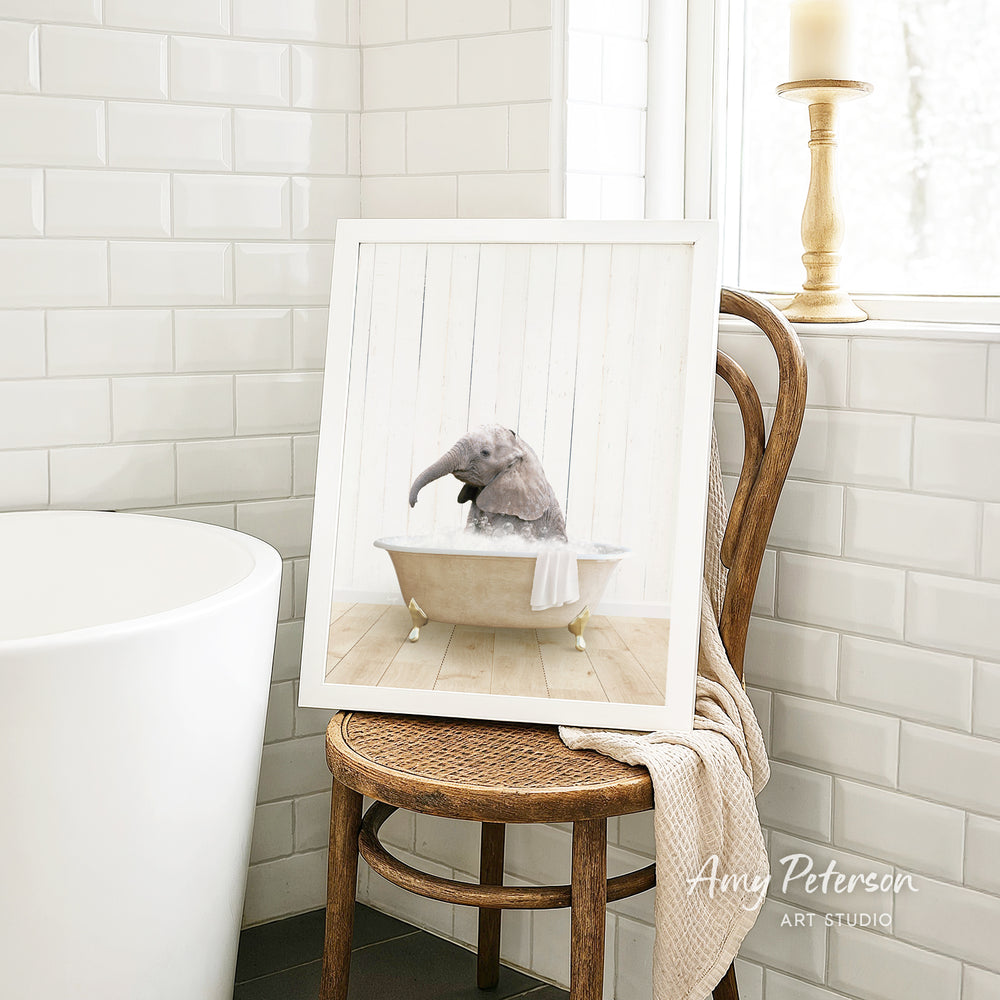 A framed image of a gray elephant sitting in a bathtub, with a towel draped over the side of the tub, is displayed on a wooden chair in a bathroom setting.