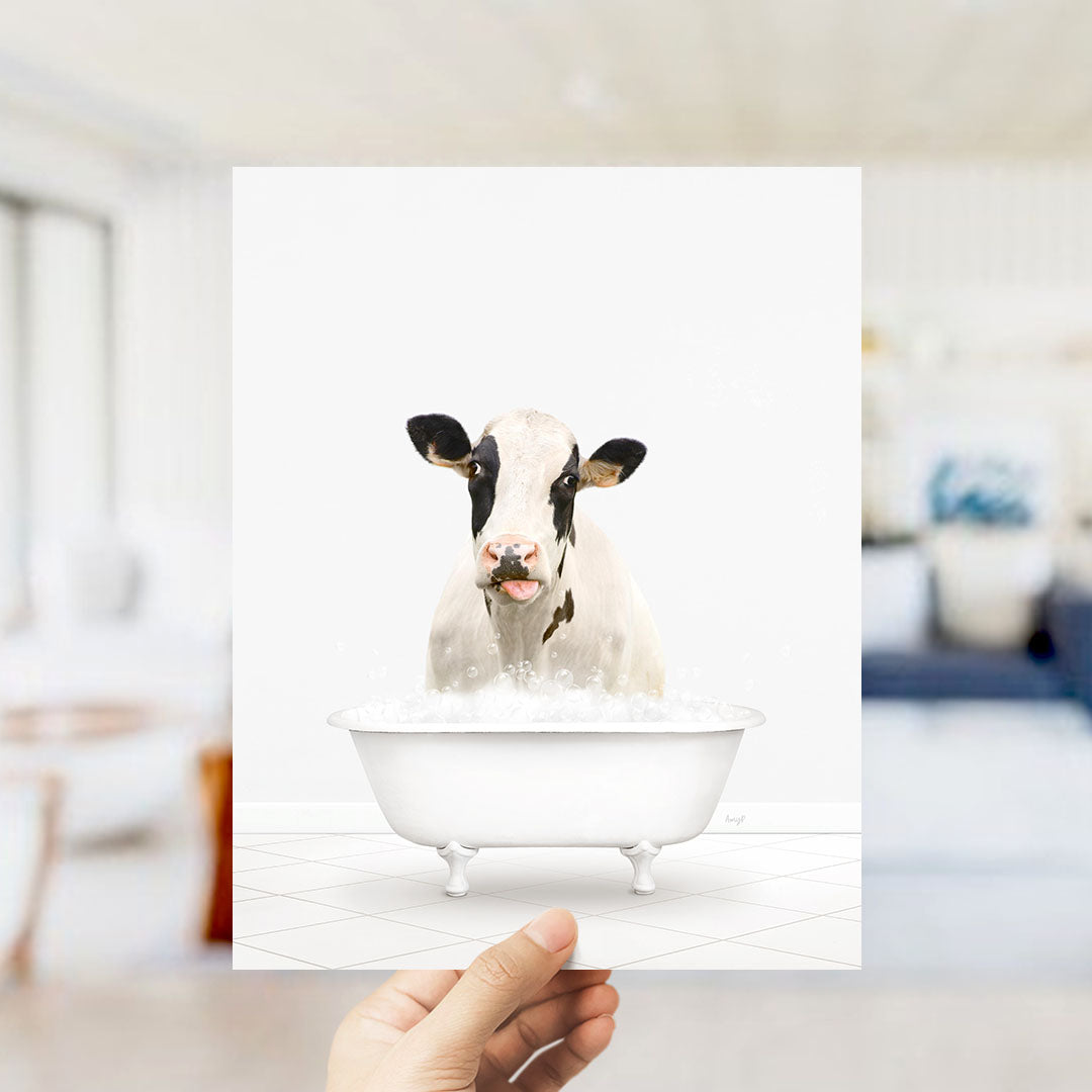 A hand holds up a framed image of a cow sitting in a bathtub filled with water.