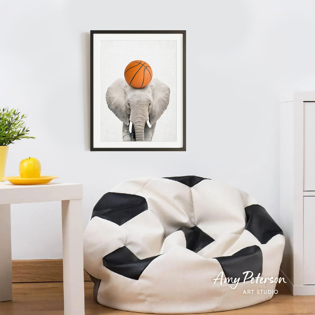 a framed picture of an elephant holding a basketball on its head, hanging on a wall in a room with a white table and a black and white bean bag chair.
