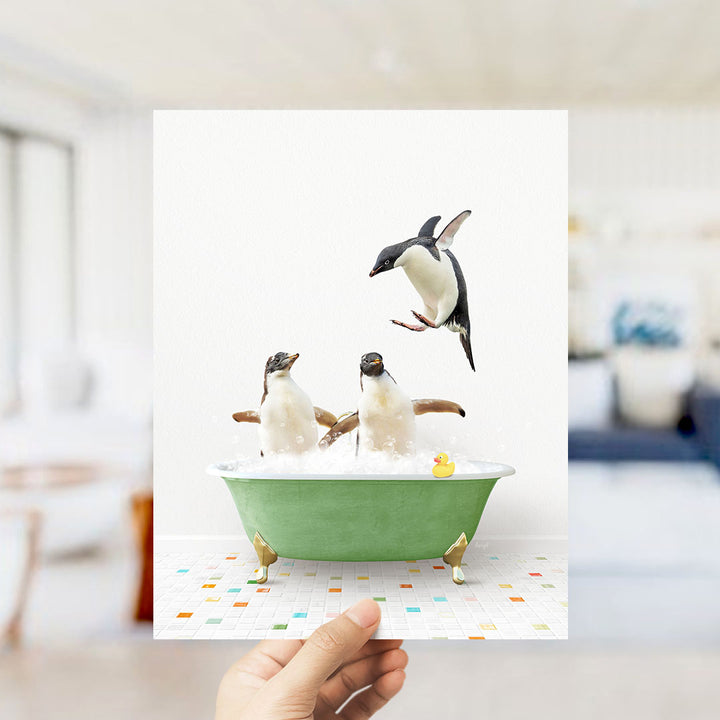 A hand holds up a framed image depicting two penguins standing in a green bathtub filled with bubbles, with one penguin flying towards the other.