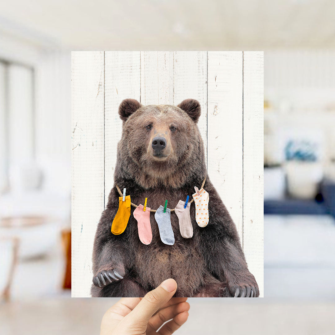 A hand holds up a framed image of a brown bear wearing colorful socks hanging on a clothesline.