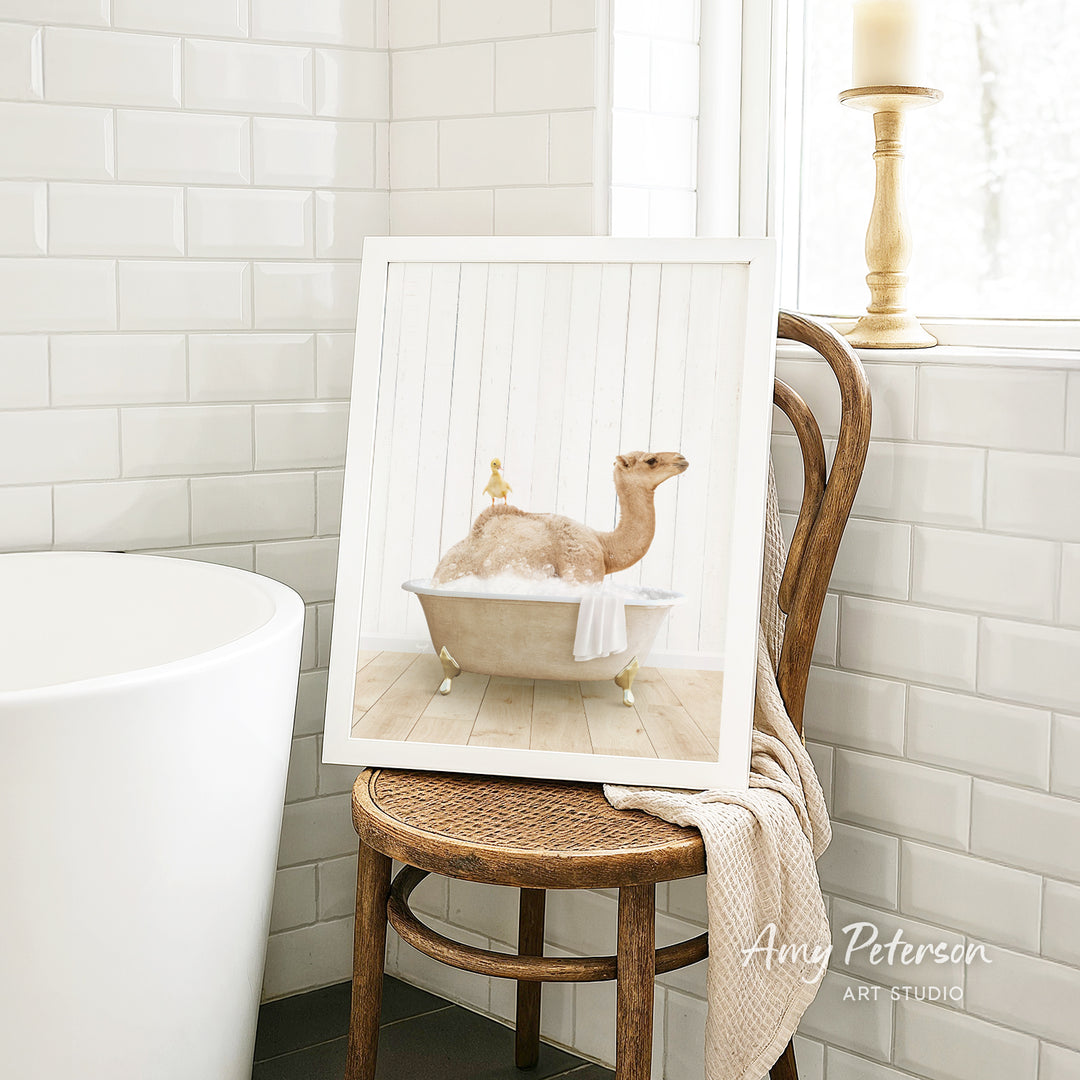 A framed image of a camel sitting in a bathtub, with a towel draped over the side of the tub, is displayed on a wooden chair in a bathroom setting.