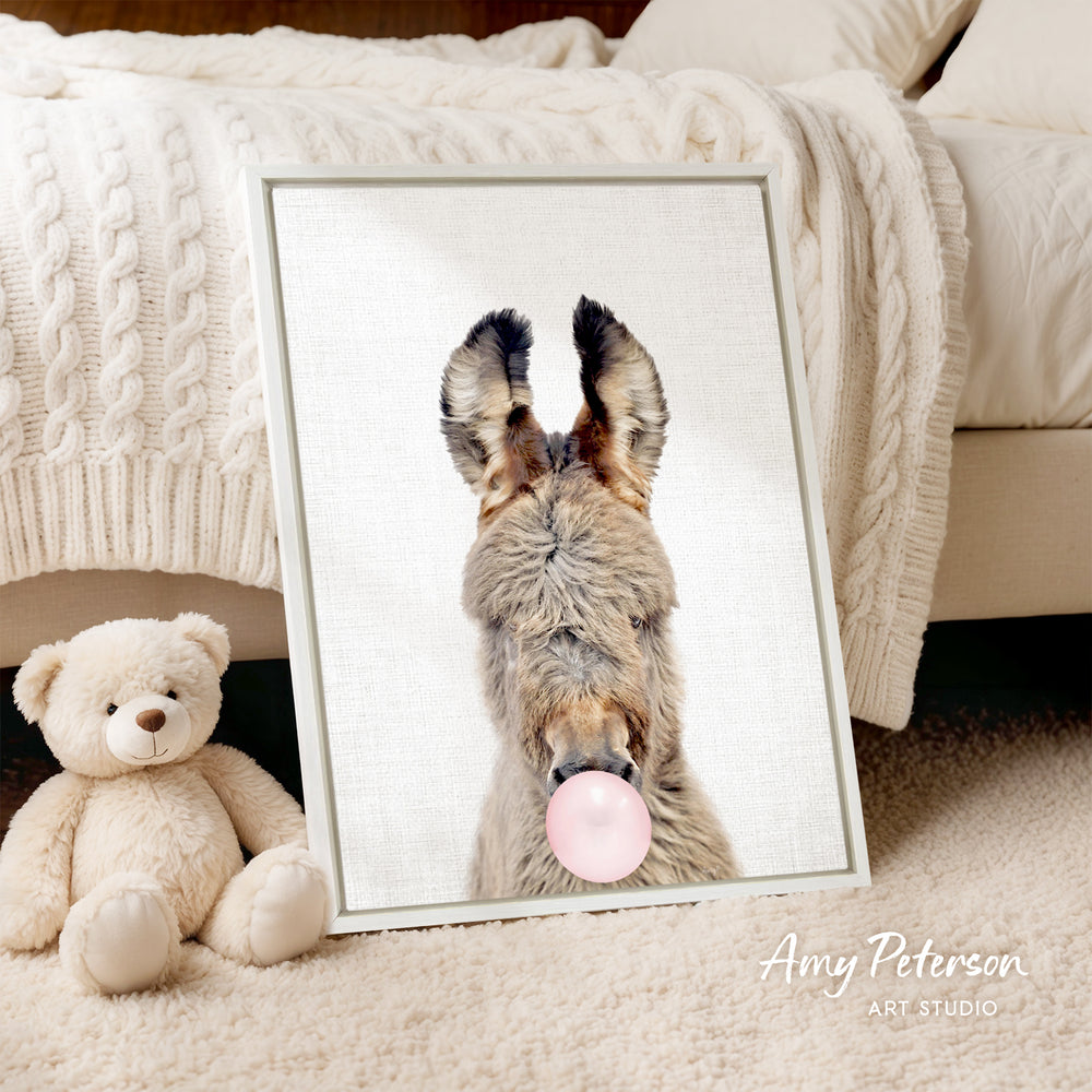 A framed image of a donkey blowing a bubble gum bubble is displayed on a white bedspread, accompanied by a teddy bear and a blanket.