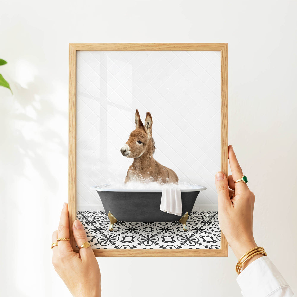 A person's hands are holding a framed picture of a donkey sitting in a bathtub filled with water and bubbles.