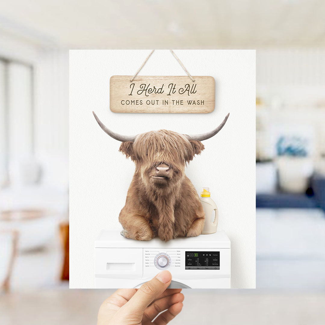 A hand holding a sign that says "I Herd It All Comes out in the Wash" above a washing machine with a large horned animal sitting on top of it.