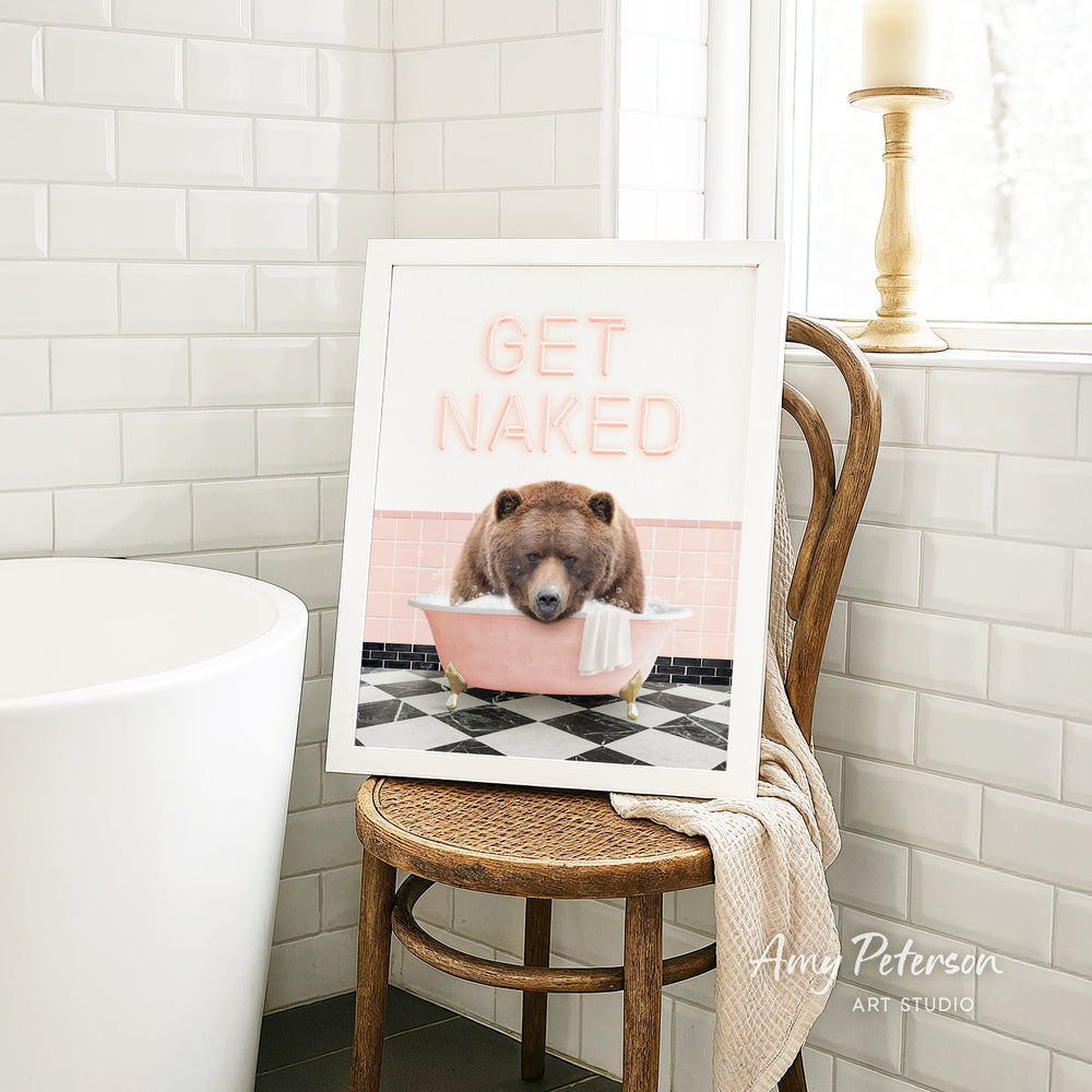 A framed image of a bear sitting in a bathtub with the text "GET NAKED" on it, placed on a wooden chair in a bathroom setting.