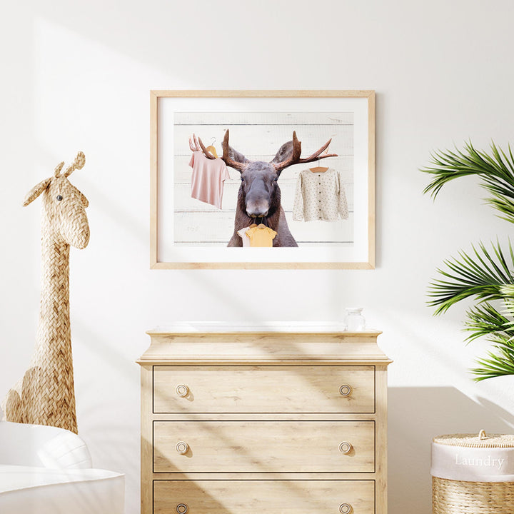 a wooden dresser with a framed picture of a moose hanging on the wall above it. The dresser has three drawers and is situated in a room with a giraffe figurine and a potted plant.