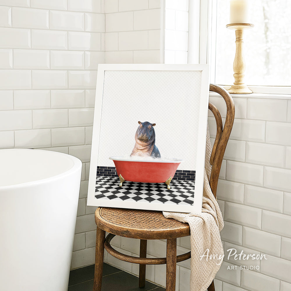 A framed image of a hippopotamus sitting in a red bathtub, surrounded by a black and white checkered floor, is displayed on a wooden chair in a bathroom setting.