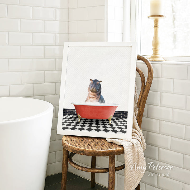 A framed image of a hippopotamus sitting in a red bathtub, surrounded by a black and white checkered floor, is displayed on a wooden chair in a bathroom setting.