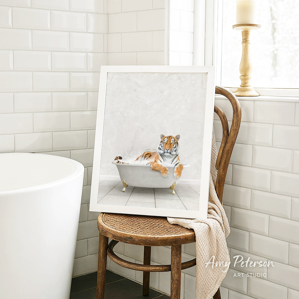 A framed painting of a tiger sitting in a bathtub is displayed on a wooden chair in a bathroom setting.