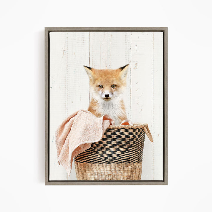 A young fox sits in a woven basket, its head resting on a pink towel.