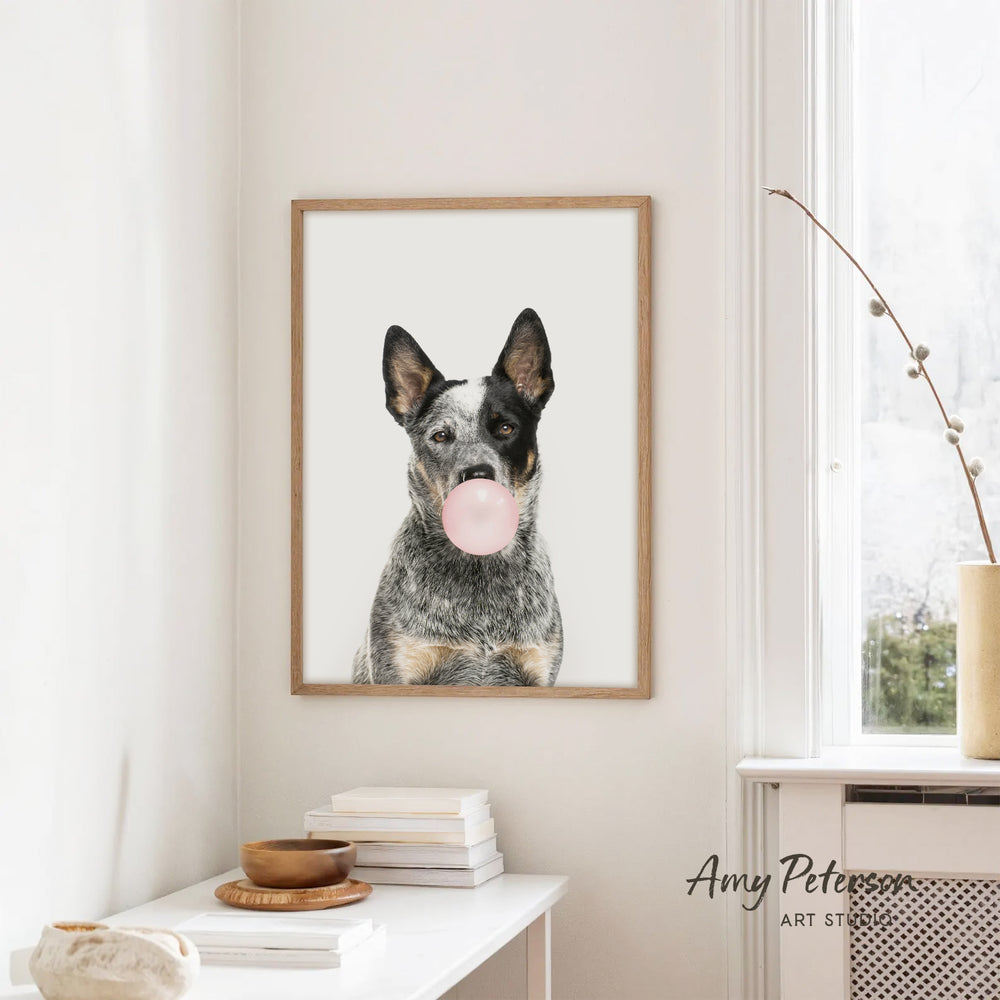 A framed image of a dog blowing a bubble gum bubble is hanging on a white wall in a room with a white table and a window.