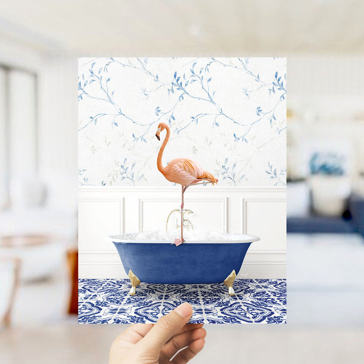 A hand holds up a framed image of a pink flamingo standing on a blue bathtub, with a floral wallpaper in the background.