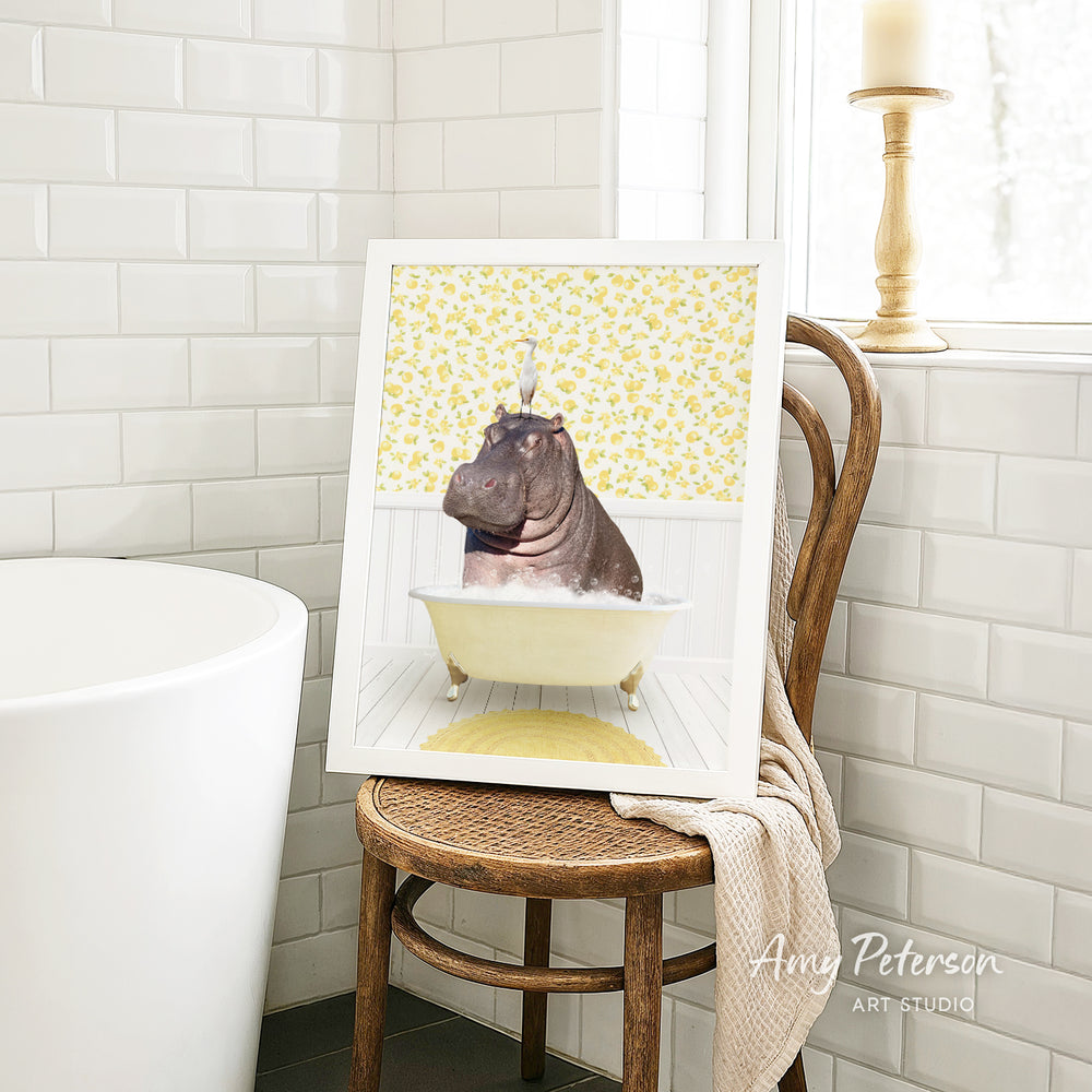 A framed image of a hippopotamus sitting in a bathtub, surrounded by yellow flowers, is displayed on a wooden chair in a bathroom setting.