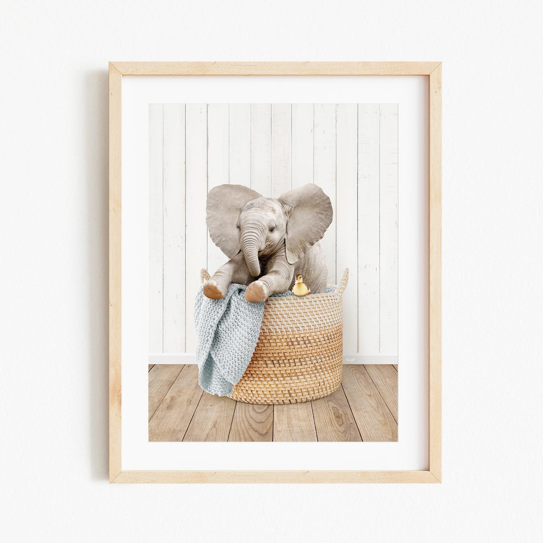 A baby elephant sitting in a woven basket, holding a blue blanket and a yellow toy.