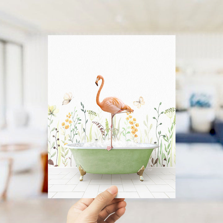 A hand holds up a framed image depicting a pink flamingo standing in a green bathtub filled with bubbles, surrounded by a floral and butterfly-themed background.
