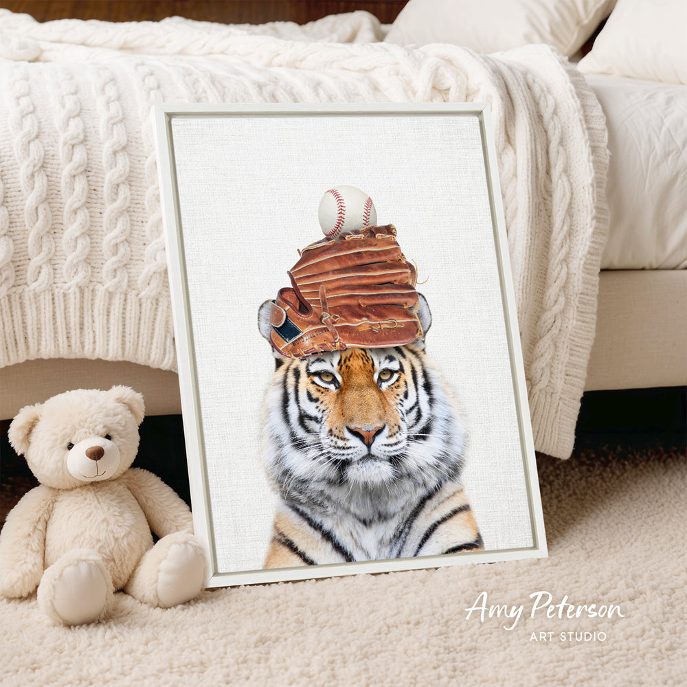 A framed print of a tiger wearing a baseball cap with a baseball and glove on top, placed on a beige carpet next to a stuffed teddy bear.