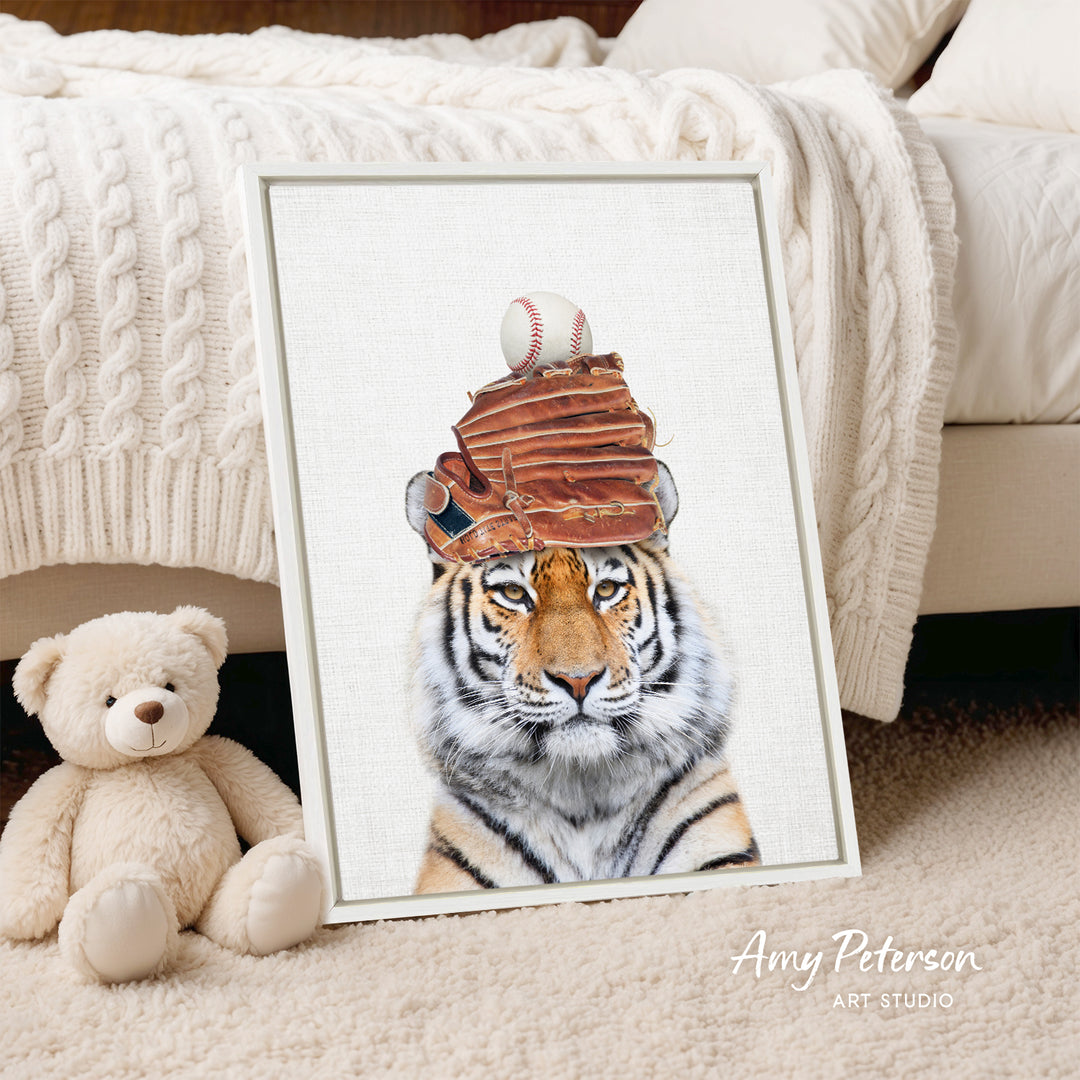 A framed print of a tiger wearing a baseball cap with a baseball and glove on top, placed on a beige carpet next to a stuffed teddy bear.
