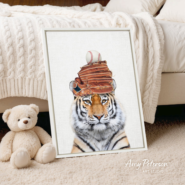 A framed print of a tiger wearing a baseball cap with a baseball and glove on top, placed on a beige carpet next to a stuffed teddy bear.