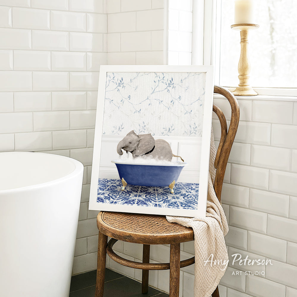 A framed image of a gray elephant sitting in a blue bathtub, surrounded by a white floral pattern, is displayed on a wooden chair in a bathroom setting.