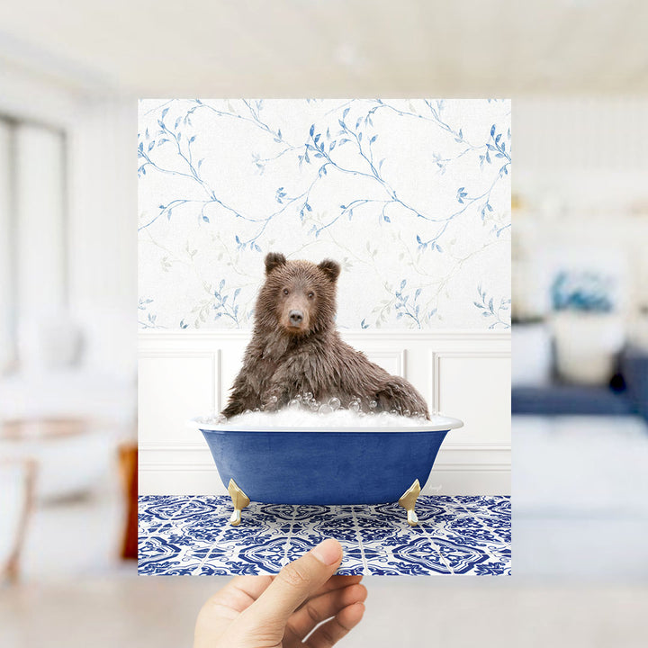 A brown bear sitting in a blue bathtub filled with bubbles, with a floral patterned wall behind it.