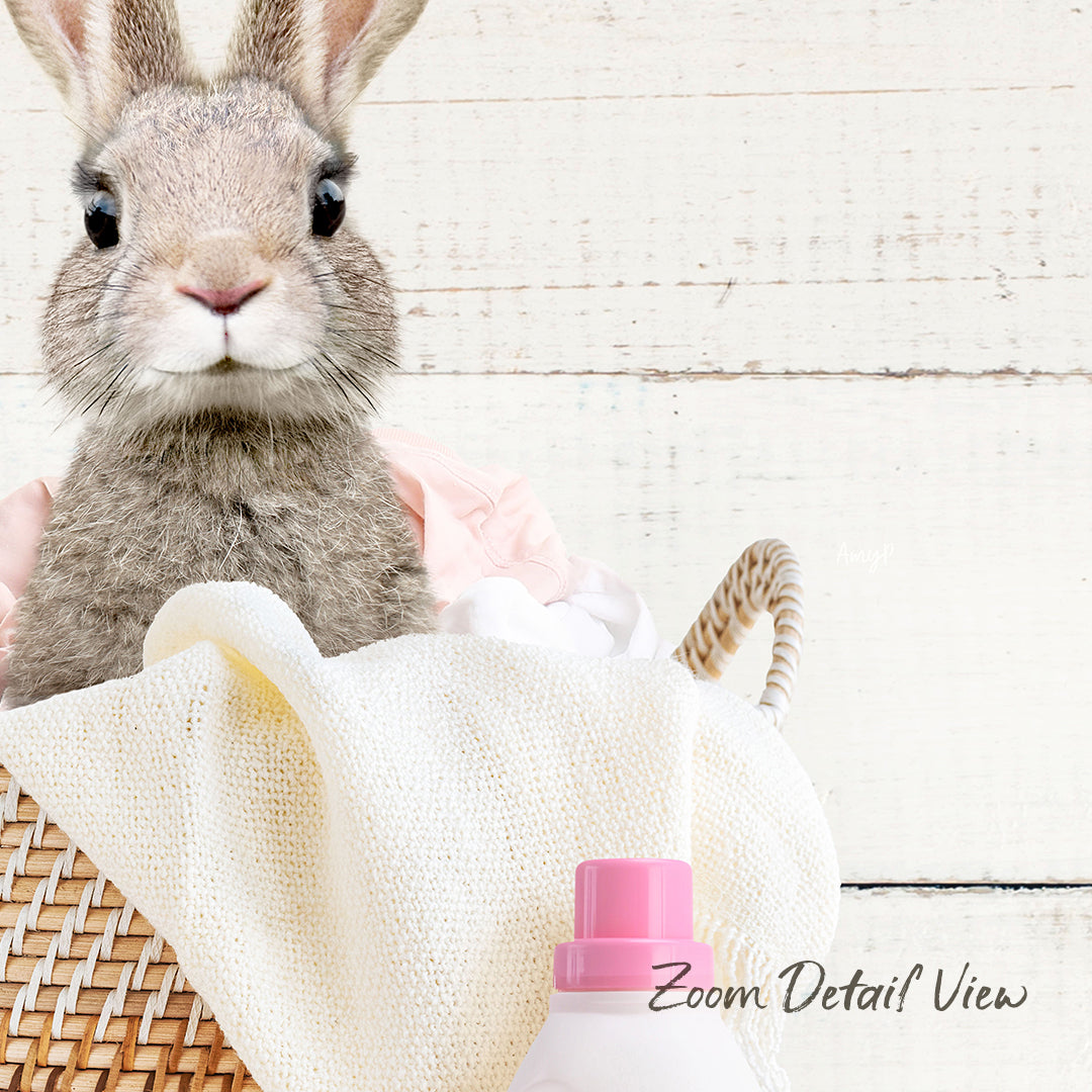 A cute gray rabbit sits in a wicker basket, surrounded by a white blanket and a pink bottle, with a wooden background.