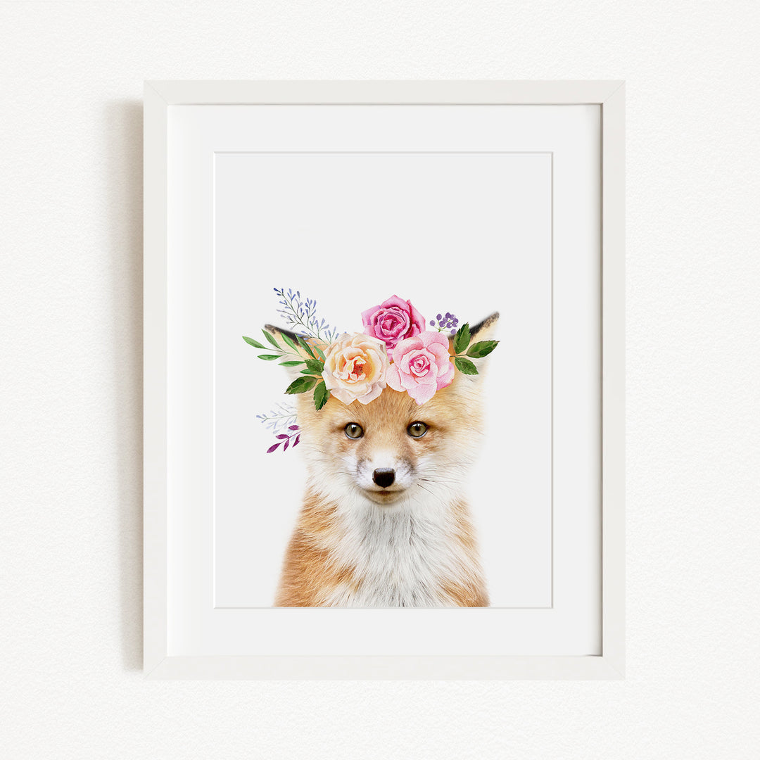 A framed print of a fox wearing a floral crown of pink and white roses and greenery.