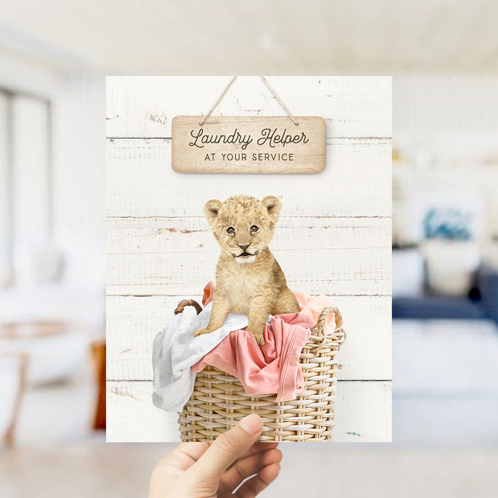 A hand holding a card with a picture of a young lion cub sitting in a wicker basket filled with clothes.
