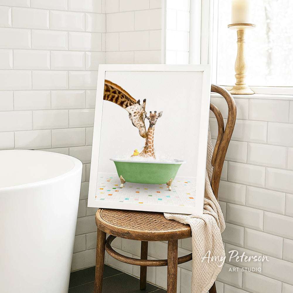 A framed image of a giraffe and a baby giraffe sitting in a green bathtub, with a towel draped over the back of a wooden chair in a bathroom setting.