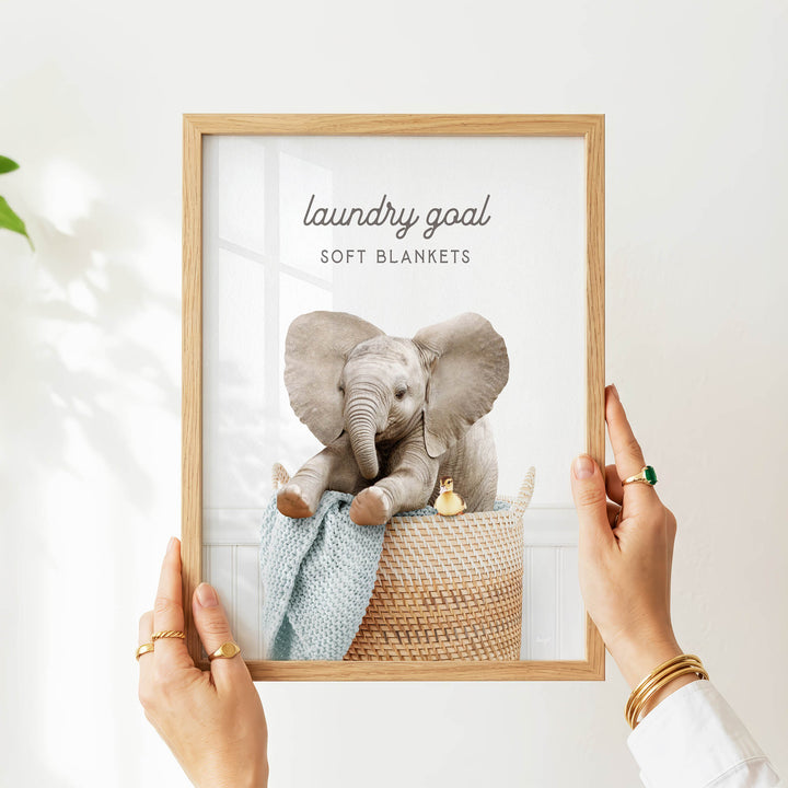 A person is holding up a framed image of a baby elephant sitting in a basket, with a blanket and a toy duck nearby.