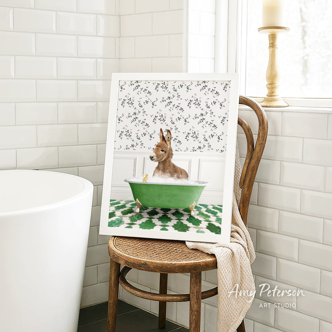 A framed image of a dog sitting in a green bathtub, surrounded by a floral pattern, is displayed on a wooden chair in a bathroom setting.