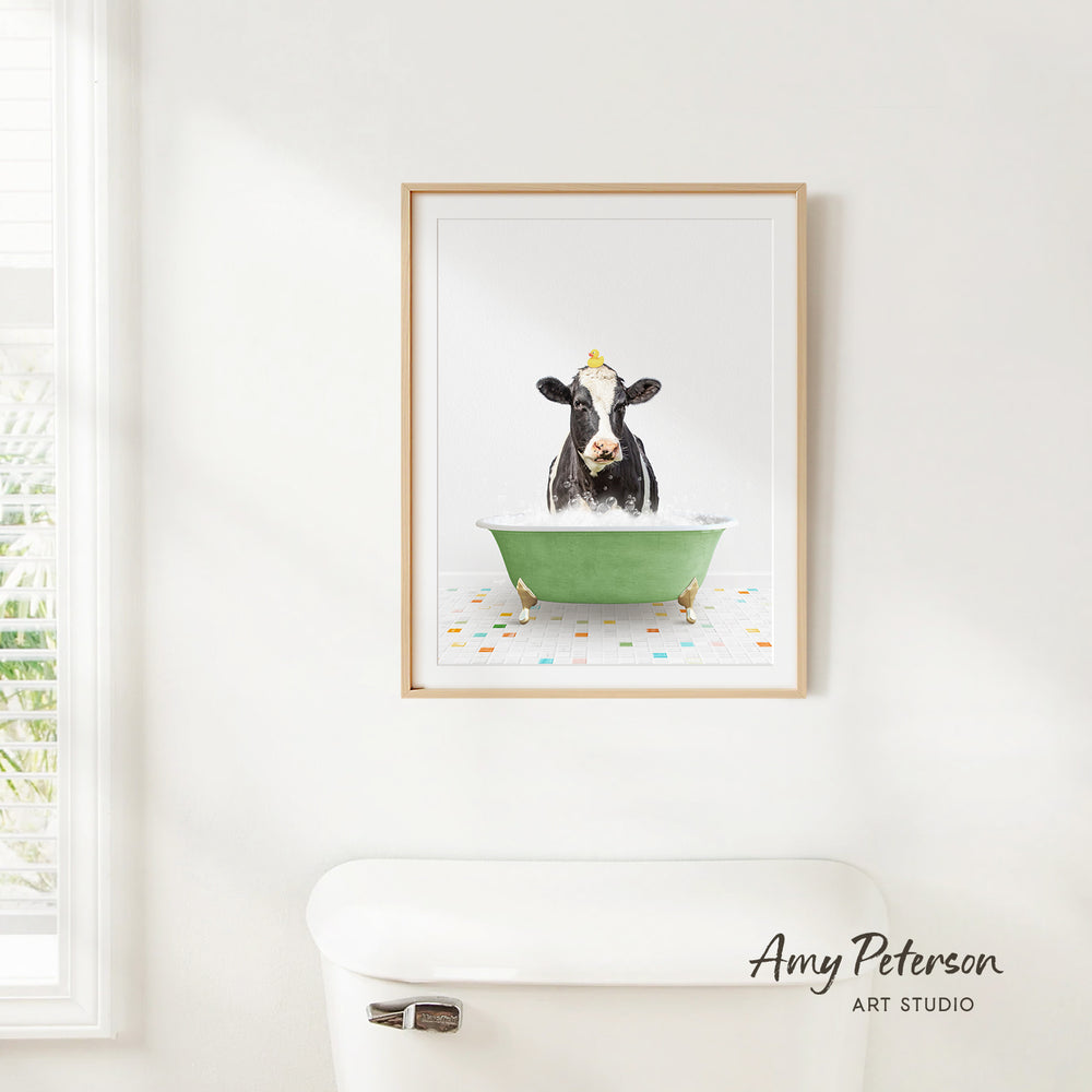 A framed print of a black and white cow sitting in a green bathtub, with a white toilet in the foreground.