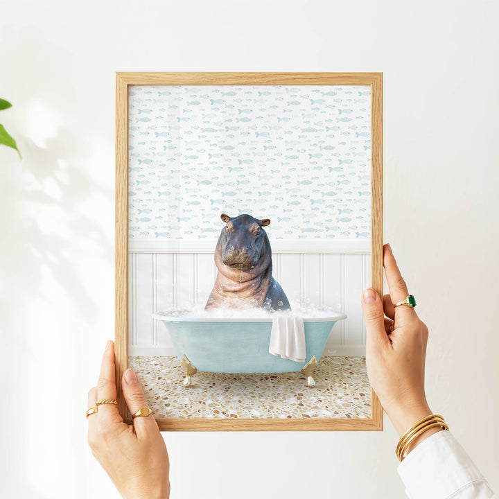 A person's hands are holding a framed picture of a hippopotamus sitting in a bathtub filled with water and bubbles.