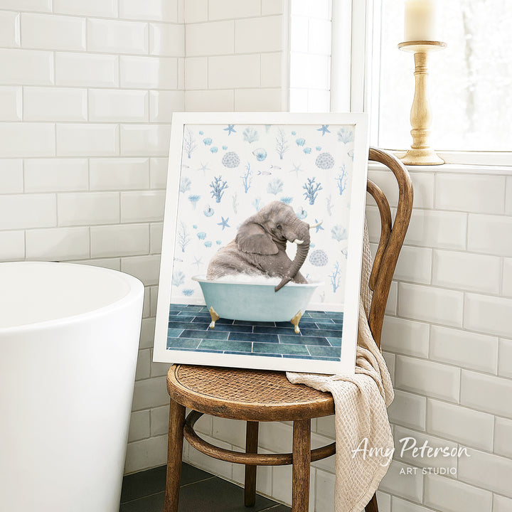A framed image of a gray elephant sitting in a blue bathtub, surrounded by blue and white floral patterns, is displayed on a wooden chair in a bathroom setting.