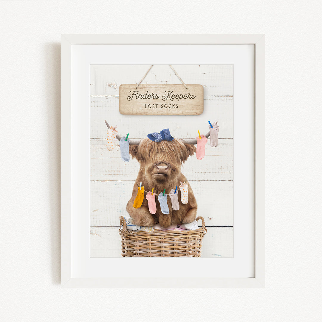 A framed image depicting a brown cow sitting in a wicker basket, with a sign above it that reads "Finders Keepers Lost Socks".