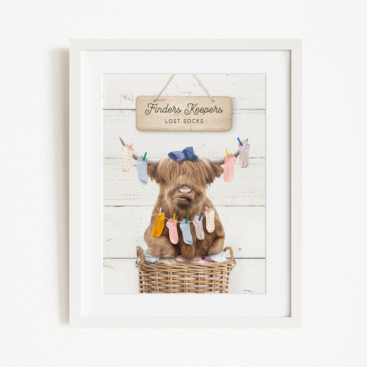 A framed image depicting a brown cow sitting in a wicker basket, with a sign above it that reads "Finders Keepers Lost Socks".