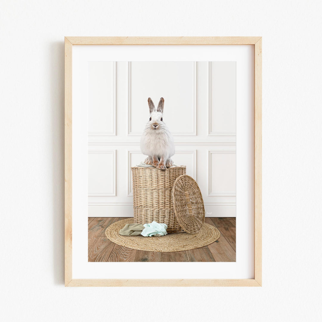 A white rabbit sits atop a woven basket on a wooden floor, with a green blanket and a basket lid nearby.