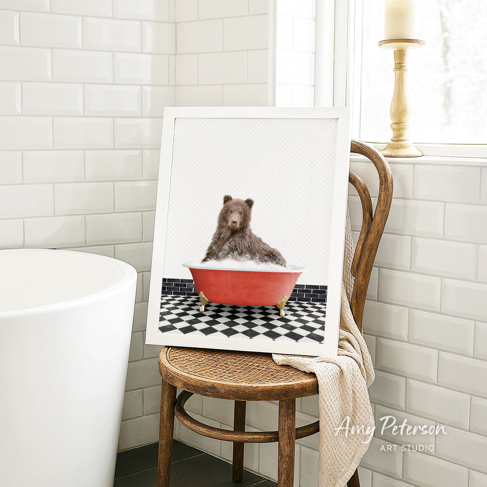 A framed image of a bear sitting in a red bathtub, surrounded by a black and white checkered floor, is displayed on a wooden chair in a bathroom setting.