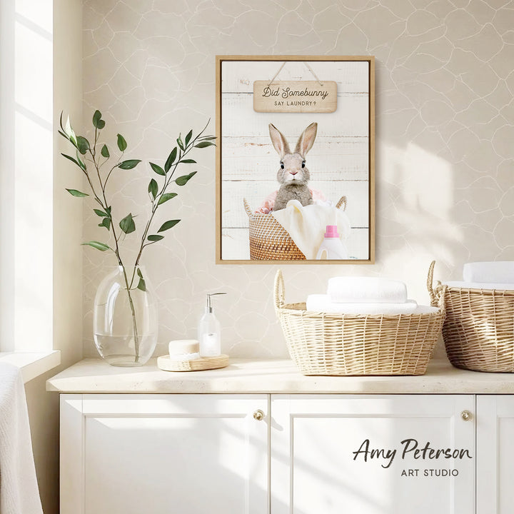 a bathroom scene with a framed picture of a rabbit in a basket, a vase with green leaves, and baskets on a counter.